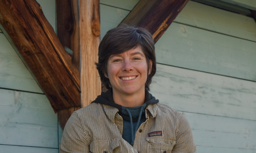 A person with short brown hair is smiling while leaning against a wooden structure. They are wearing a striped jacket over a dark hoodie. The background features blue painted wooden panels.